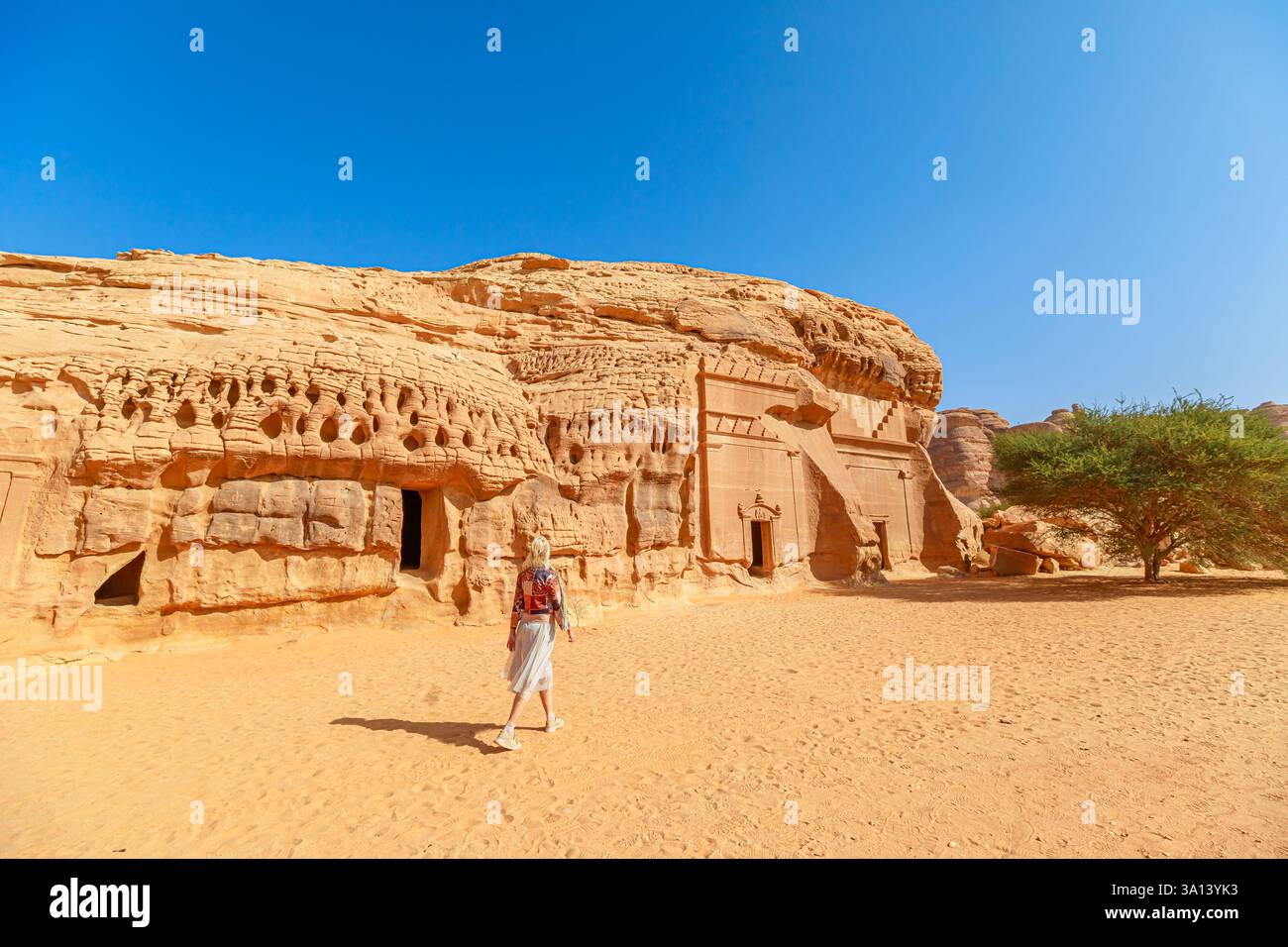 Touriste blonde marchant dans le désert à côté de Jabal AlBanat et tombeau géant taillé dans la roche du Royaume Nabatéen dans le site archéologique de Hegra Madain Banque D'Images