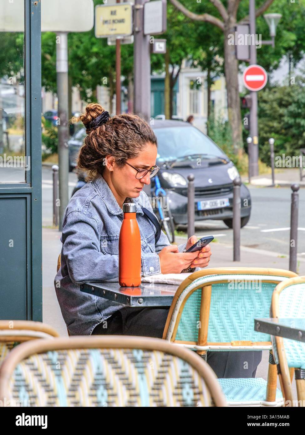 Jeune femme assise à la table de terrasse extérieure du café regardant smartphone -Montparnasse-Raspail, Paris 75014, France. Banque D'Images