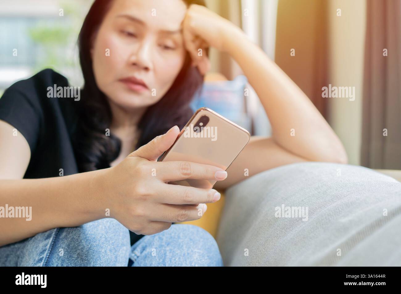 Femme asiatique adulte se sentant ennuyé ou triste tout en jouant smartphone dans le salon à la maison. Concept de vie. Mise au point sélective. Banque D'Images