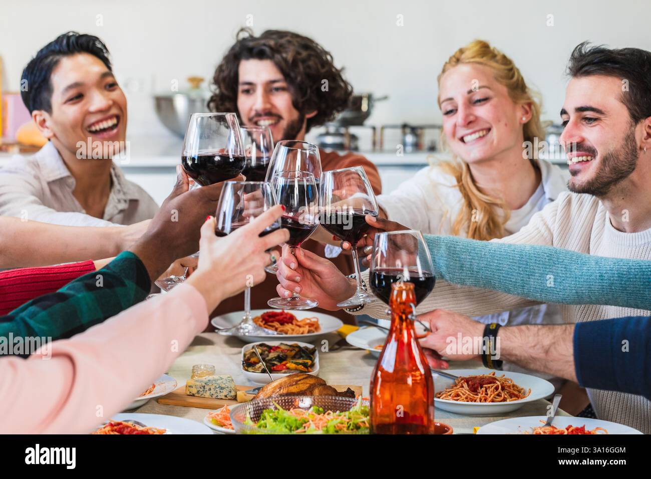 Des amis multiethniques joyeux grillent des verres de vin rouge tout en dégustant un dîner italien à la maison Banque D'Images