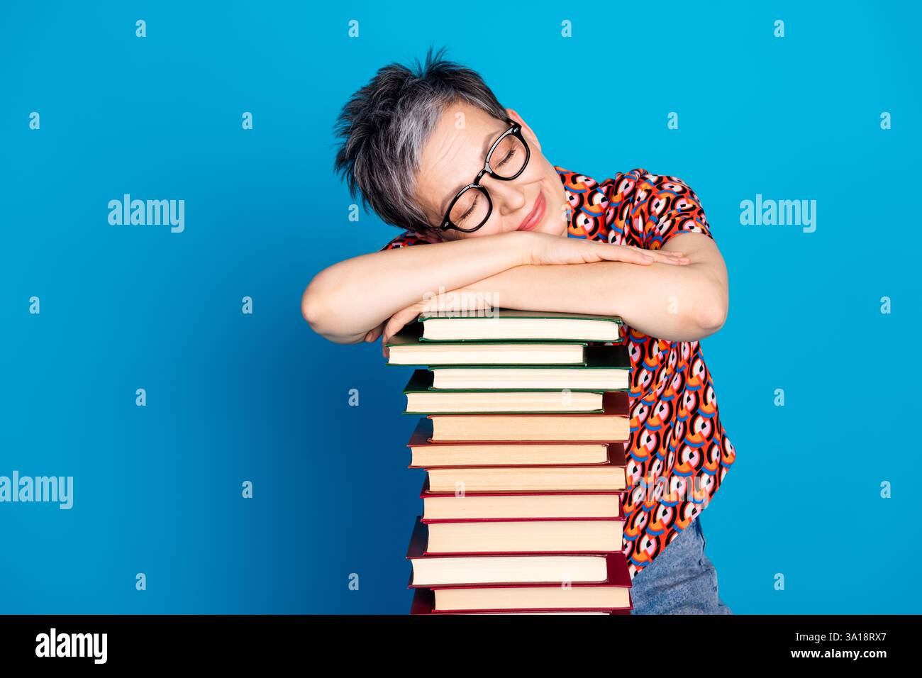 Femme âgée souriante appuyée sur une pile de livres, portant des lunettes et une chemise vibrante sur un fond bleu Banque D'Images