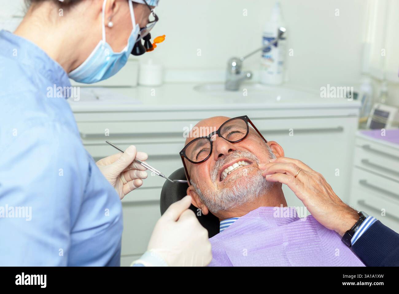 Dentiste portant une loupe et un masque chirurgical examinant un patient âgé souffrant de maux de dents dans une clinique dentaire brillante Banque D'Images