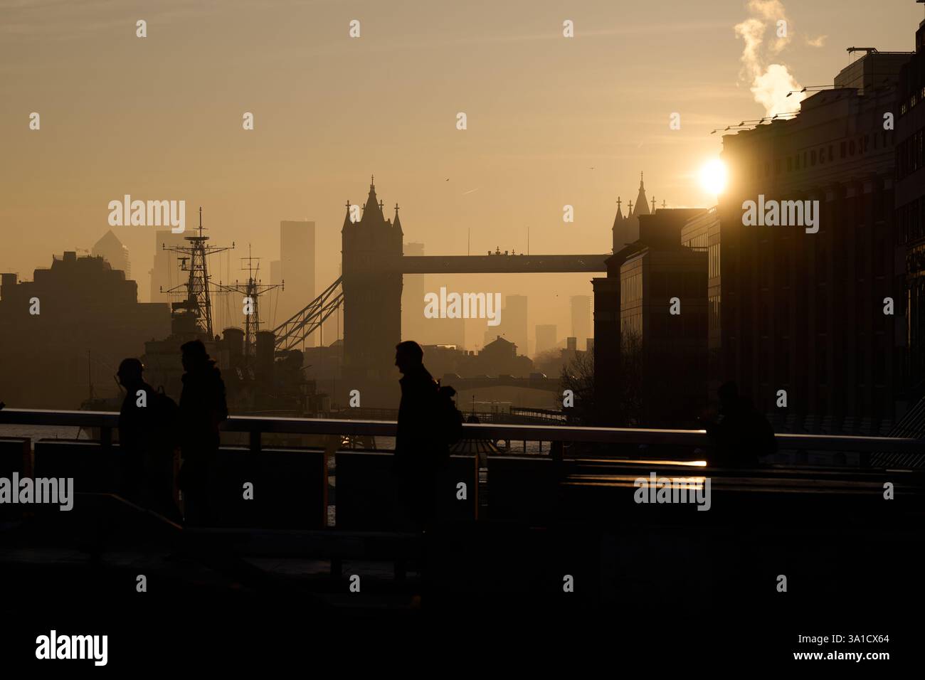 Vue tôt le matin des navetteurs marchant sur London Bridge avec vue sur Tower Bridge et HMS Belfast silhouetté par le soleil tôt le matin. Banque D'Images