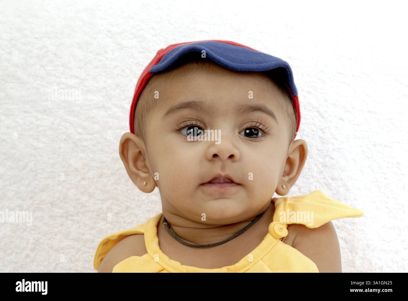 Bébé indien sud-asiatique, avec une expression coquine, côté regardant, blouse de couleur jaune, chapeau de couleur rouge, fond blanc, Inde MR#152 Banque D'Images