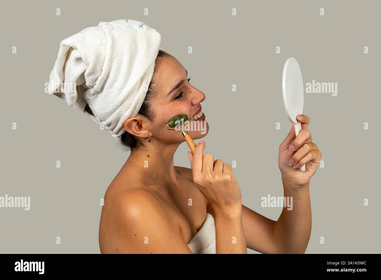 Femme heureuse utilisant le masseur à rouleaux de jade sur le visage et regardant dans le miroir après la douche, appréciant le traitement de beauté Banque D'Images