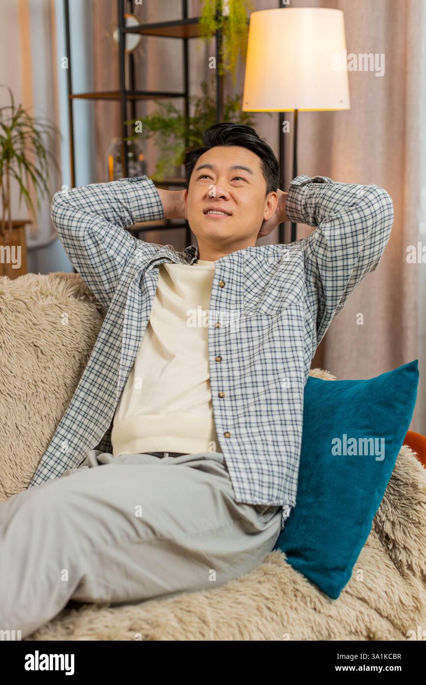 Heureux homme chinois d'âge moyen insouciant assis confortablement avec la main derrière la tête sur le canapé dans le salon à la maison. Souriant mec coréen se relaxant sur le canapé pensant regarder autour dans la chambre de l'appartement. Banque D'Images