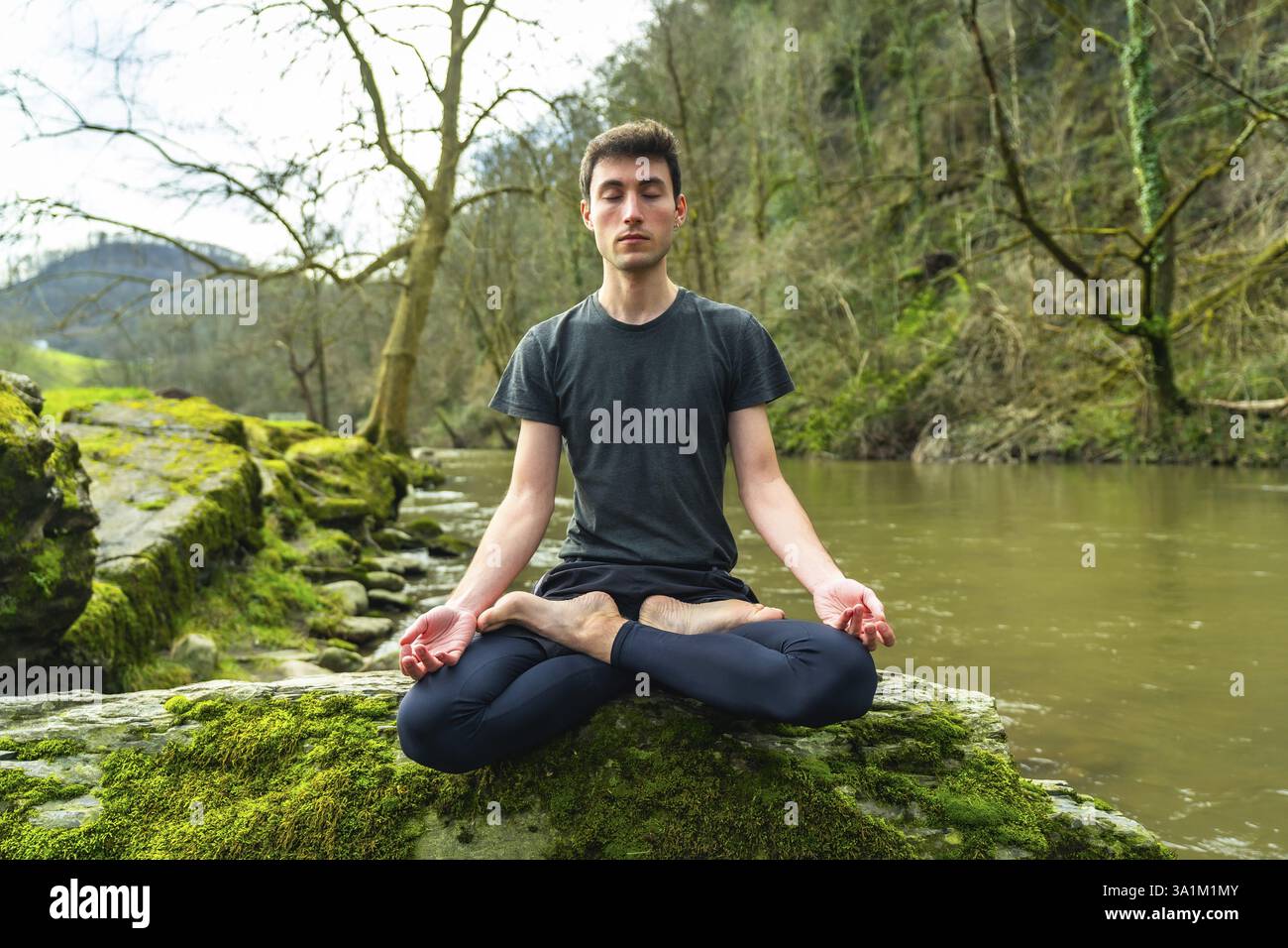 Professeur de yoga méditant en position lotus sur un rocher couvert de mousse par une rivière dans un environnement naturel paisible Banque D'Images