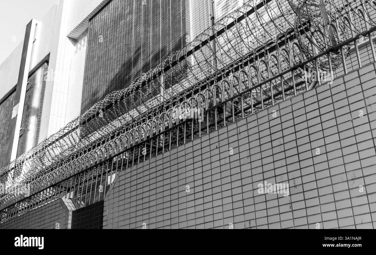 Un bâtiment sécurisé avec de hauts murs, une clôture métallique accrochée et des fils de corruption en noir et blanc. Banque D'Images