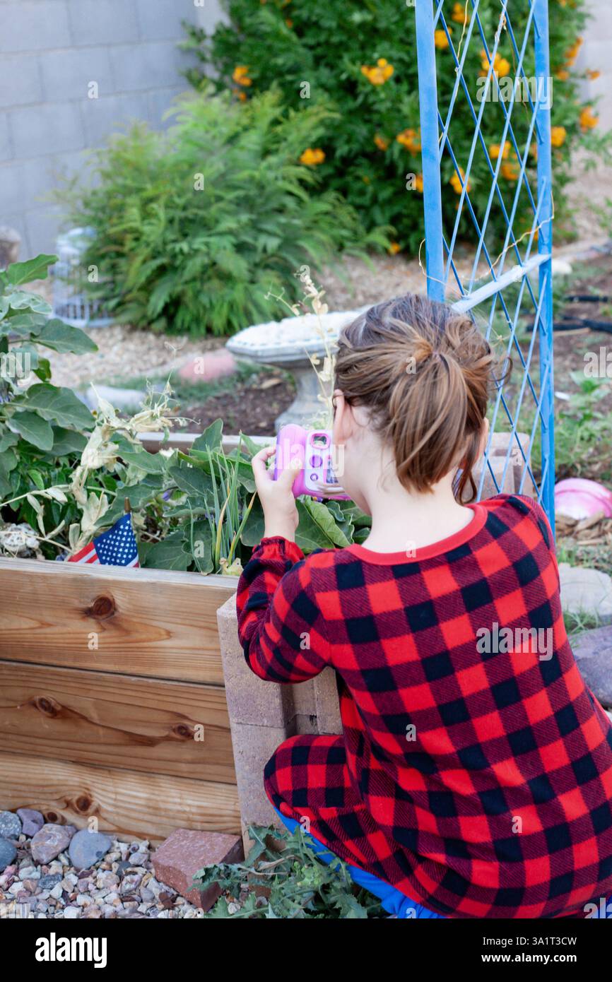 Enfant en plaid rouge capturant des plantes de jardin avec un appareil photo jouet rose Banque D'Images
