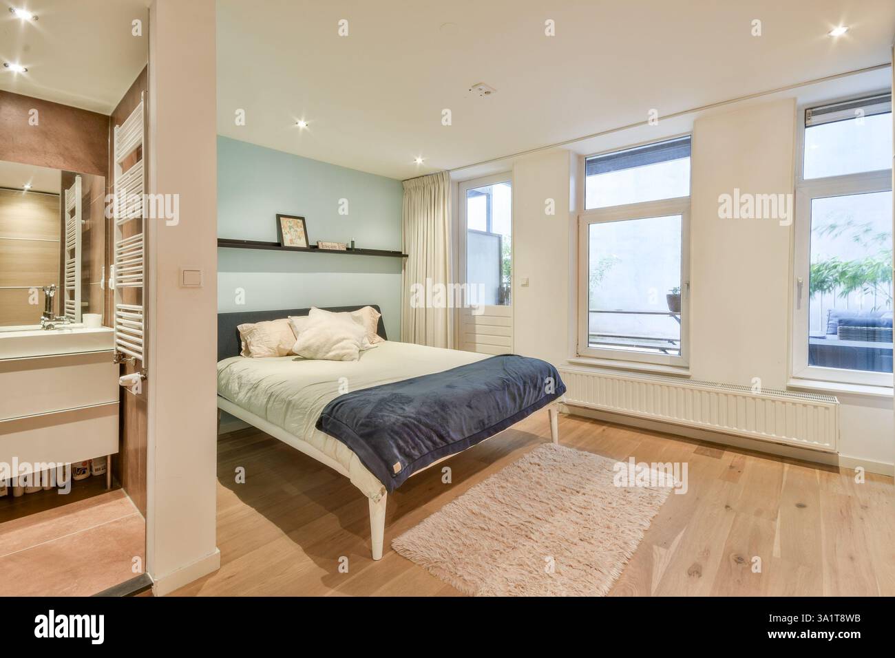 Une chambre contemporaine avec un lit confortable, du parquet clair et une lumière naturelle abondante qui traverse de grandes fenêtres, créant une atmosphère sereine. Banque D'Images