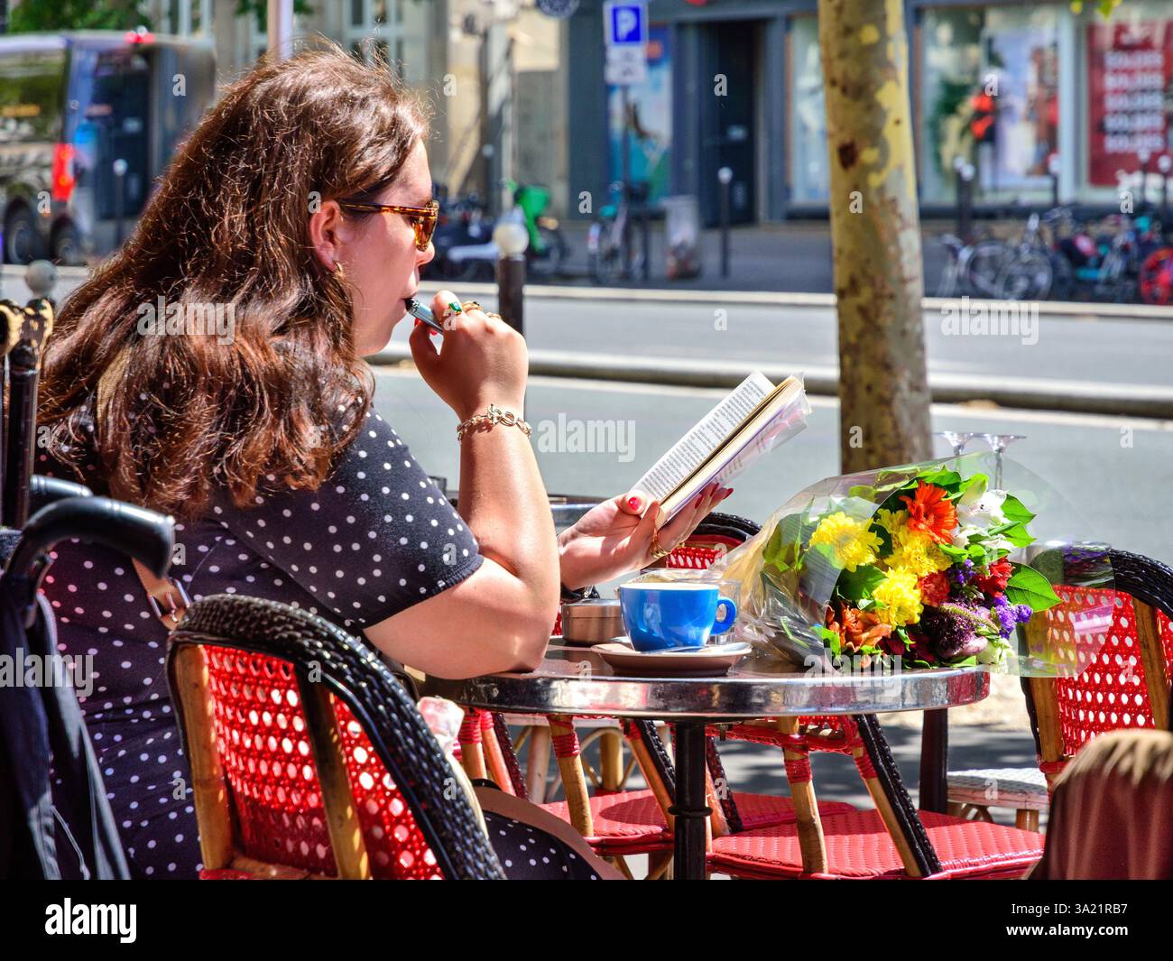 Femme assise à une table de café en plein air avec un bouquet de fleurs, vapotant et lisant un livre - Montreuil (93100), Seine-Saint-Denis, France. Banque D'Images