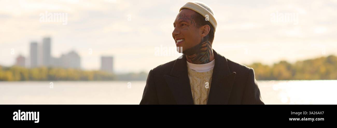Jeune homme avec des tatouages met en valeur la mode tendance tout en souriant au bord du lac paisible. Banque D'Images