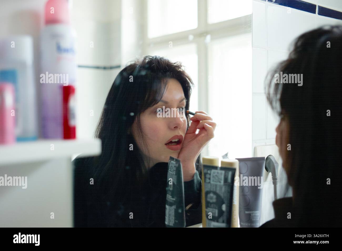 Femme appliquant le maquillage dans un miroir de salle de bain avec une expression focalisée. Berlin, Allemagne Banque D'Images