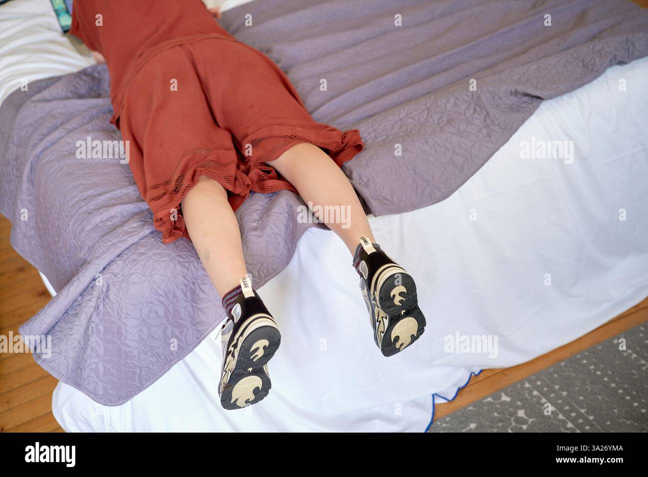 Femme en robe rouge s'écrasant face cachée sur un lit d'hôtel avec des draps violets et blancs. Hauts-de-France, France Banque D'Images