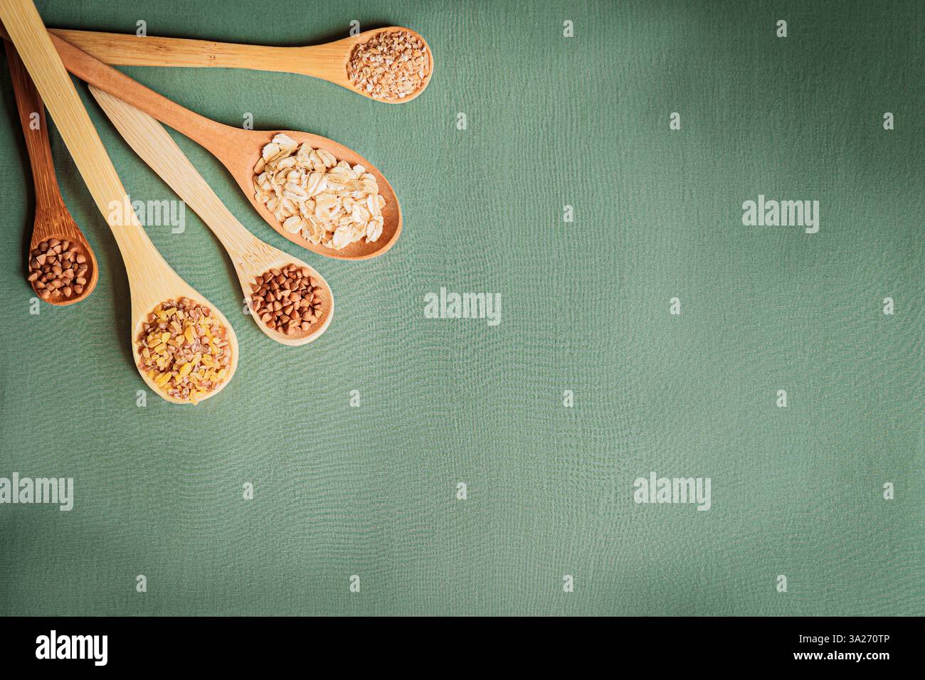 Cuillères en bois remplies d'avoine, de pois chiches, de sarrasin et de grains mélangés étalés en éventail dans un motif circulaire sur un fond vert. Composition équilibrée wi Banque D'Images
