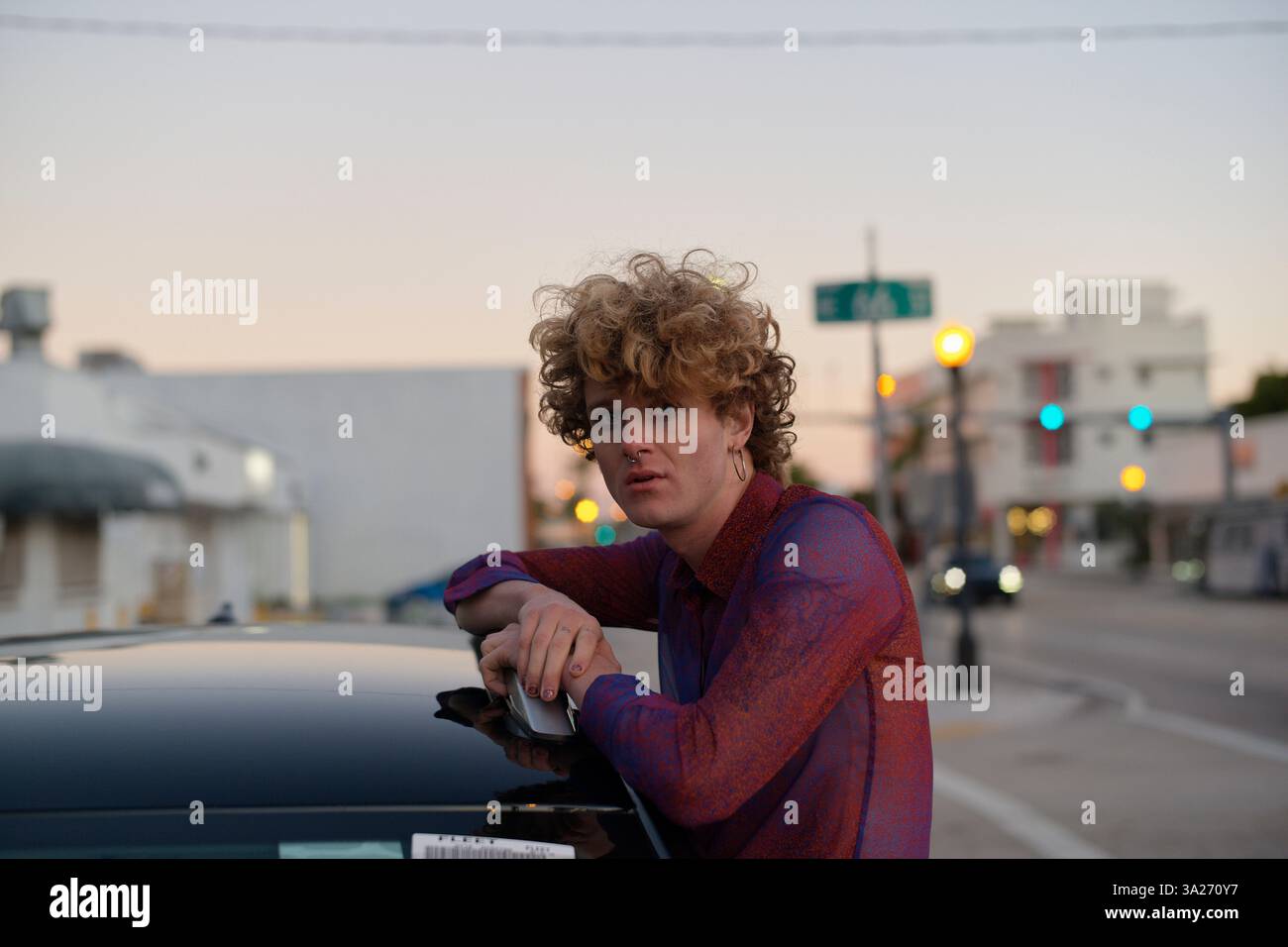 Jeune homme aux cheveux bouclés appuyé sur une voiture dans une rue de la ville au coucher du soleil. Miami, Floride, États-Unis Banque D'Images