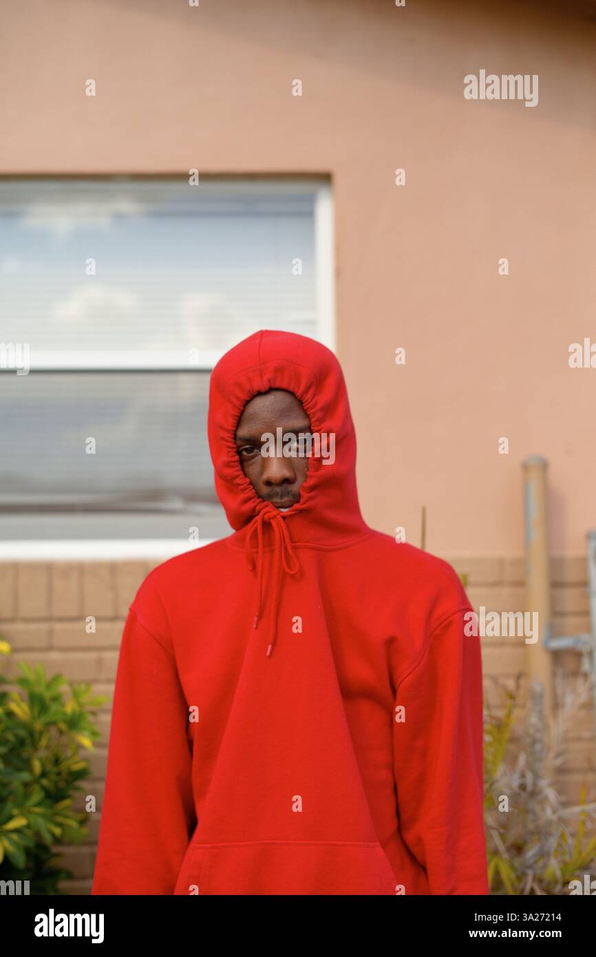 Jeune homme dans un sweat à capuche rouge se tient devant une fenêtre sur un mur beige avec des plantes à proximité. Miami, Floride Banque D'Images