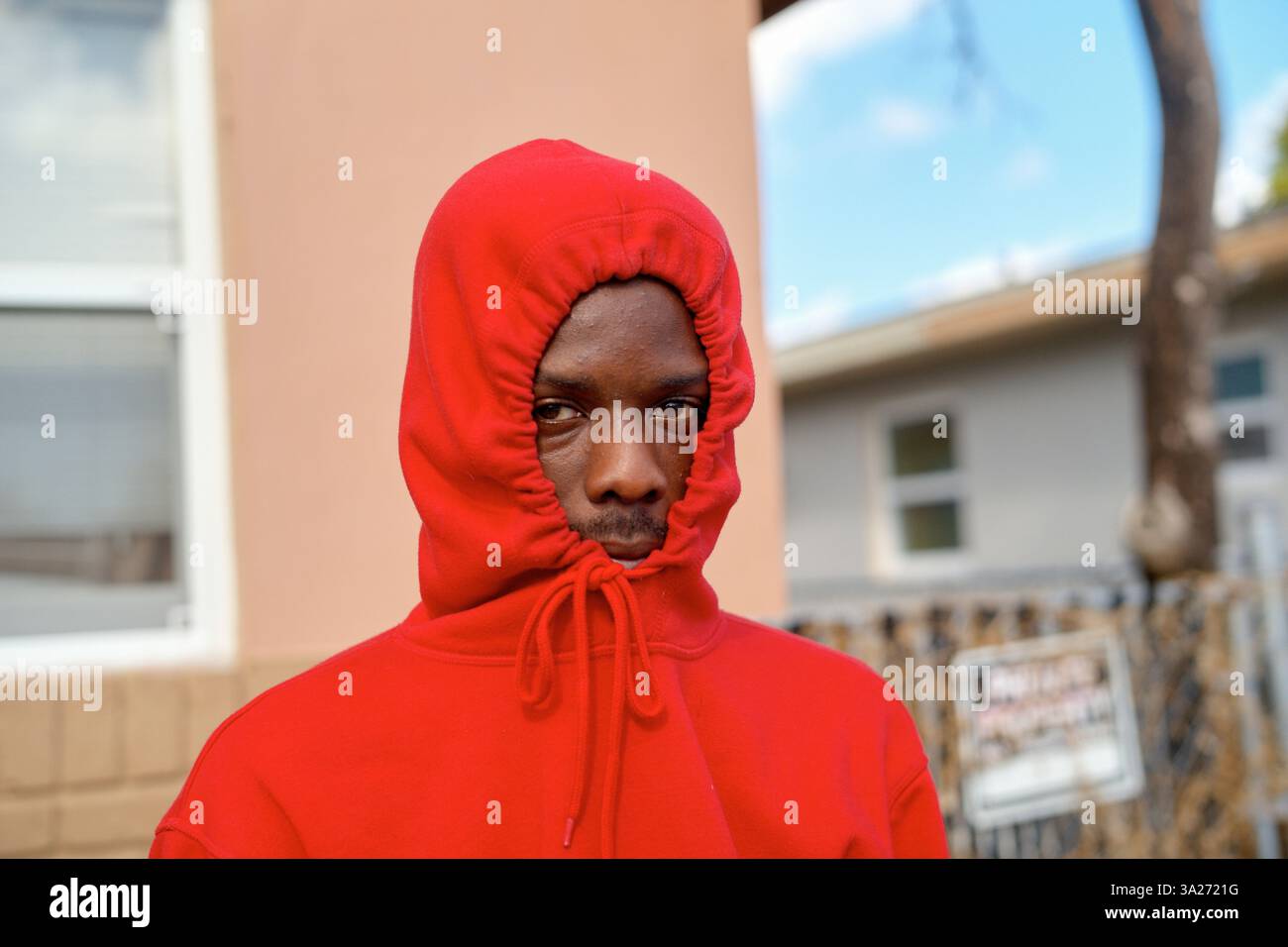 Jeune homme dans un sweat à capuche rouge se tient à l'extérieur dans une zone de banlieue, visage principalement couvert, regardant la caméra. Miami, Floride Banque D'Images