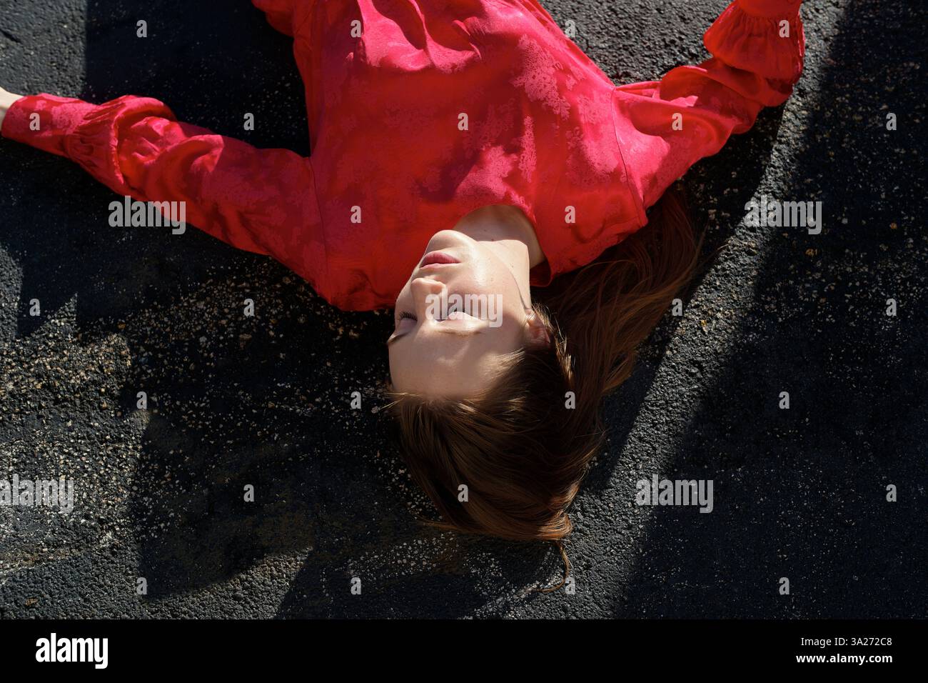 Femme en robe rouge couchée sur l'asphalte avec la lumière du soleil projetant des ombres sur son visage Miami, FL, USA Banque D'Images