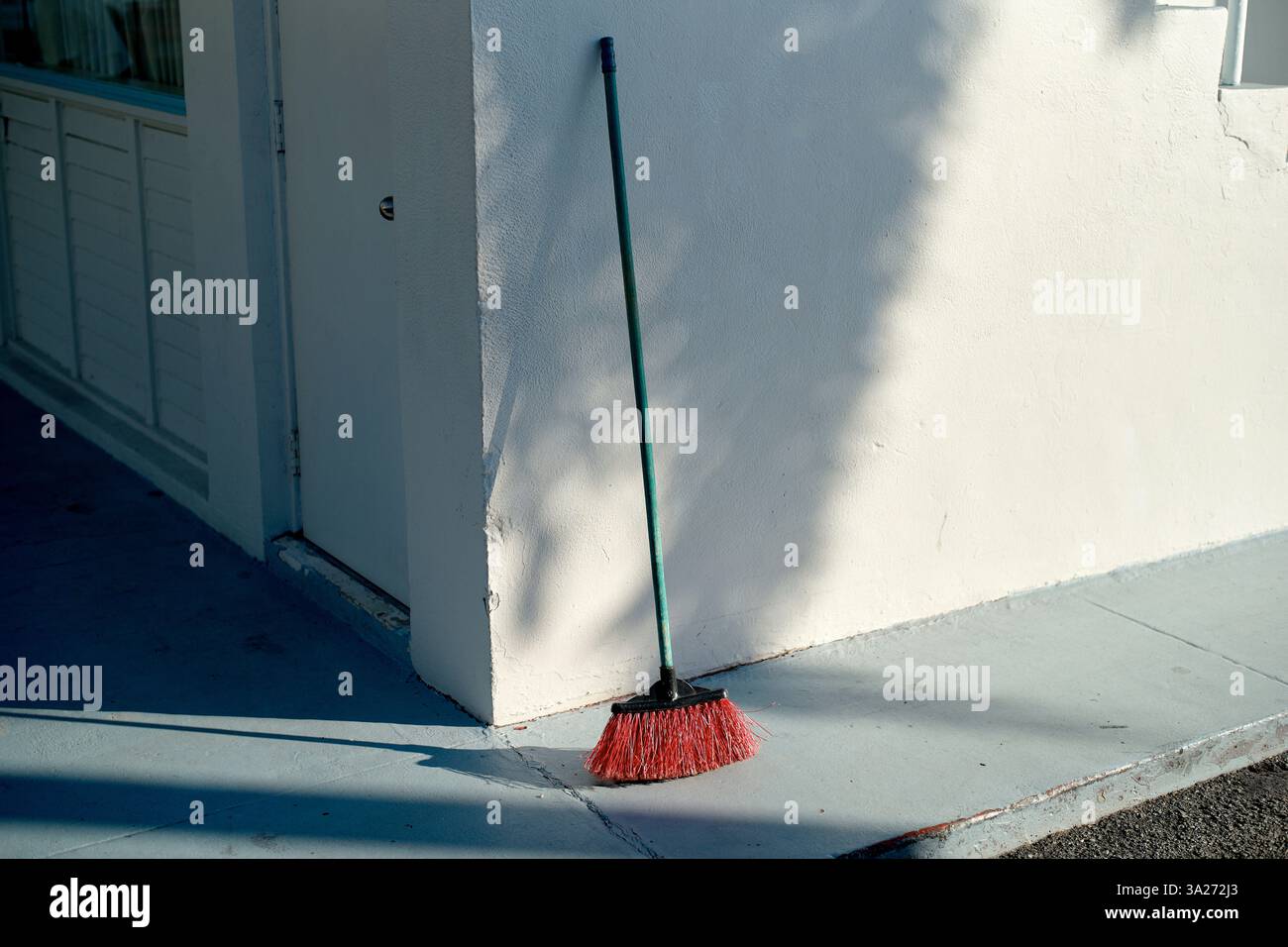 Un balai à poils rouges s'appuie contre un mur blanc, projetant une ombre sur un trottoir en ciment. Miami, Floride, États-Unis Banque D'Images