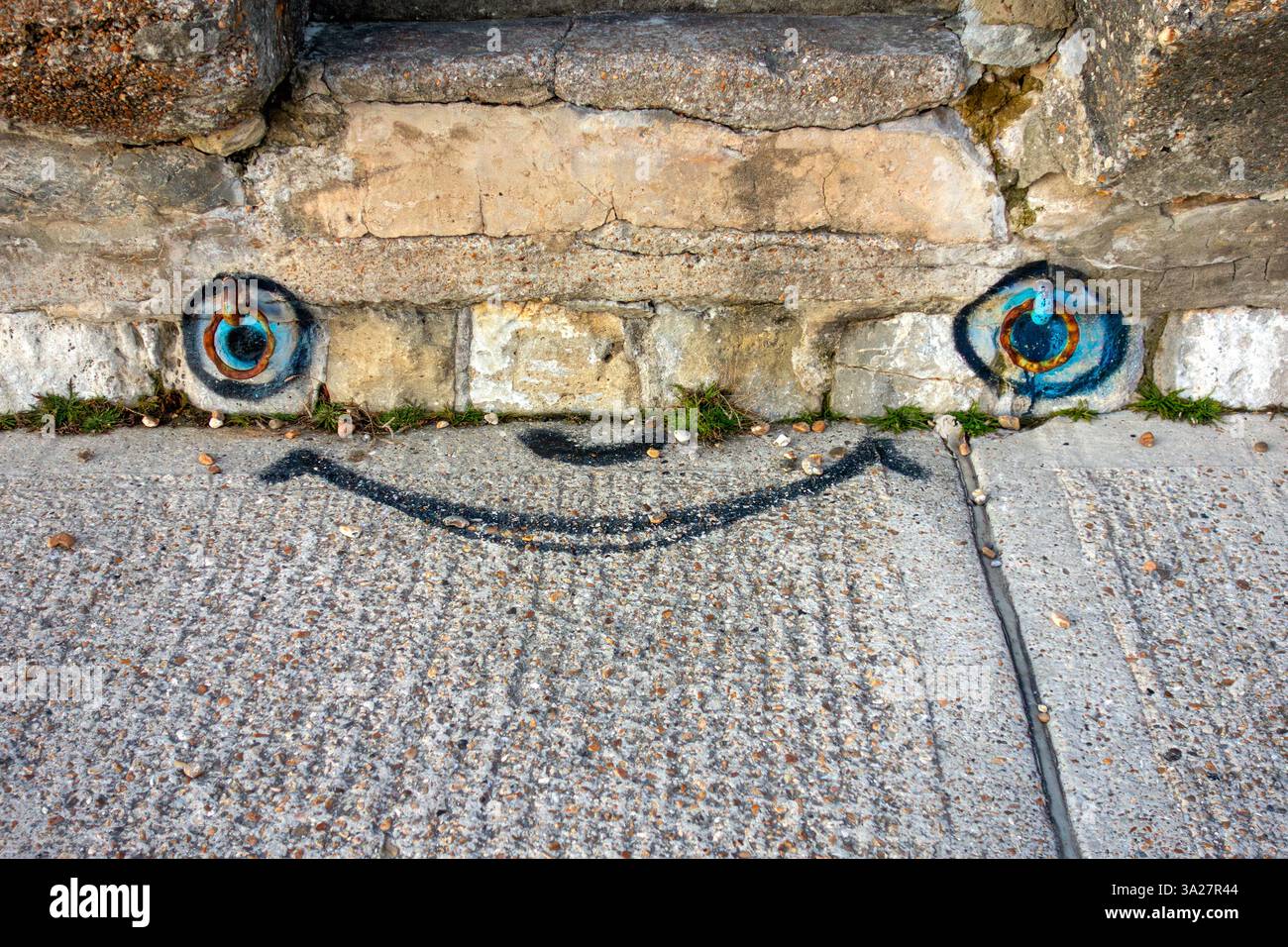 Un visage souriant dessiné sur un chemin en béton, avec des yeux positionnés autour de vieux anneaux d'amarrage rouillés Banque D'Images