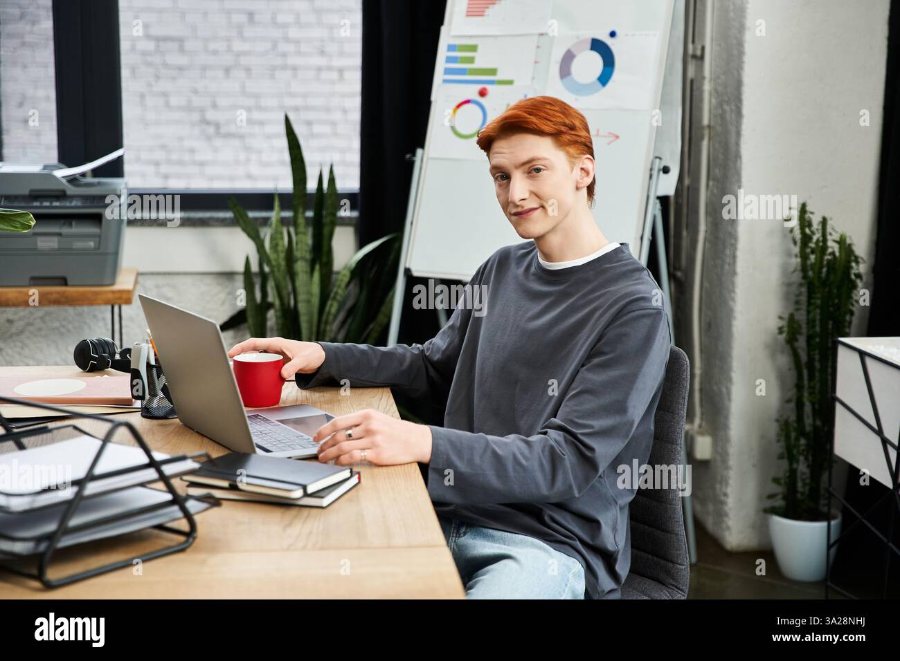 Un jeune homme aux cheveux roux s'engage activement avec son ordinateur portable dans une configuration de bureau contemporaine. Banque D'Images