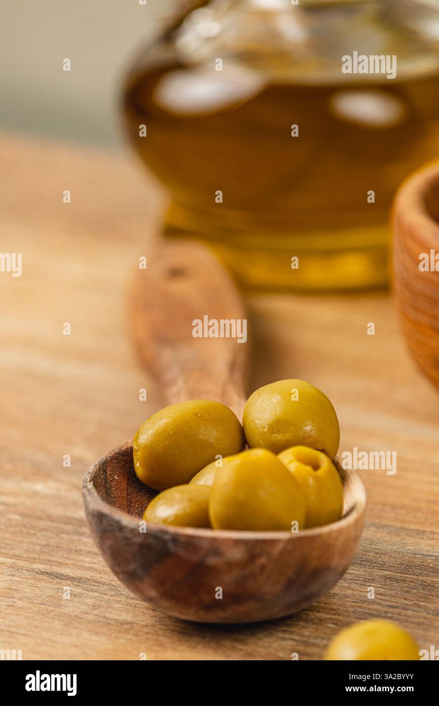 Les olives vertes fraîches reposent dans un bol en bois sculpté, mettant en valeur leur couleur vibrante. Une bouteille d'huile d'olive brille en arrière-plan, mettant en valeur cette culina Banque D'Images