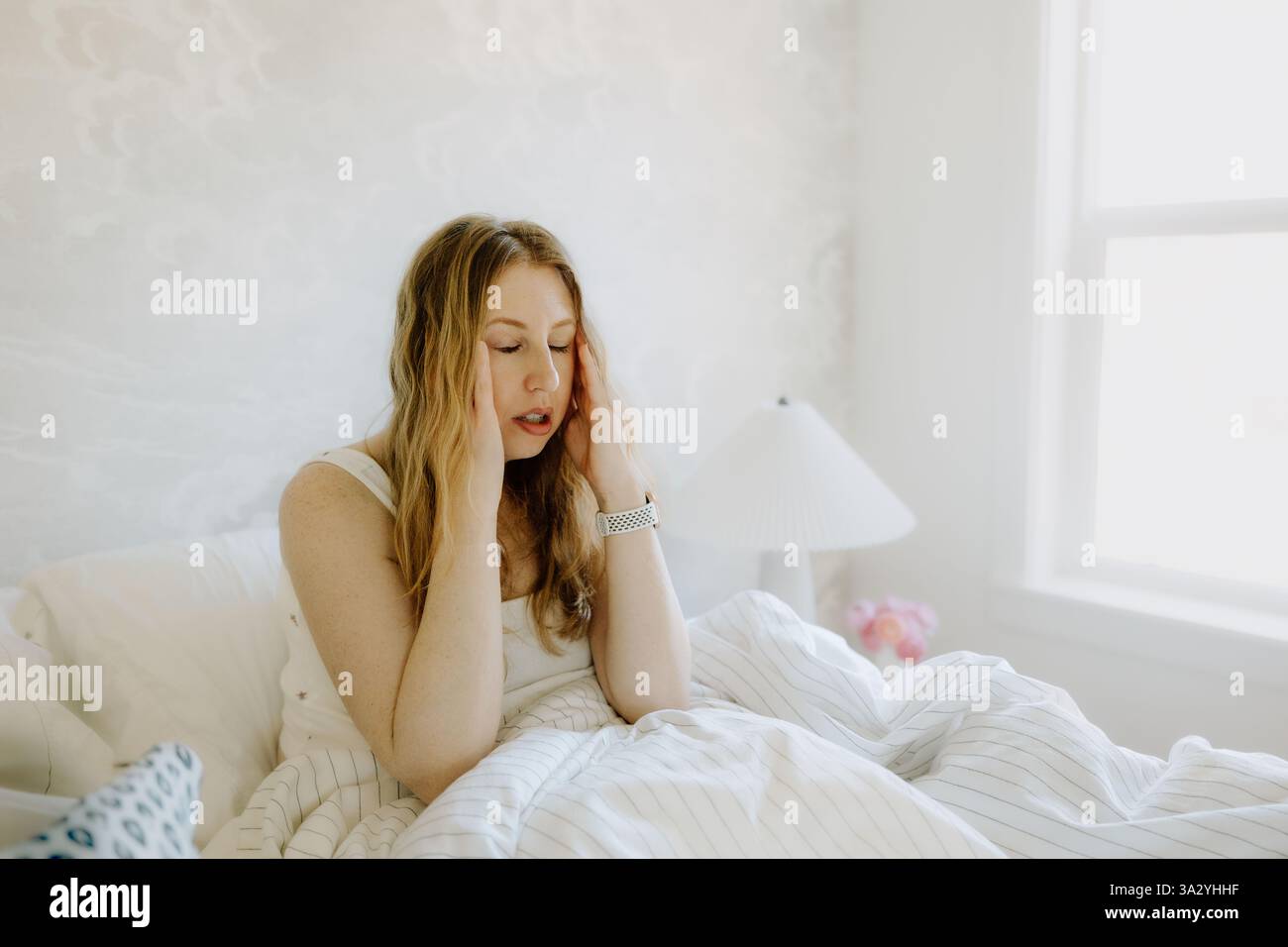 Une femme dans les années 40 est assise dans son lit, tenant sa tête dans un moment de stress Banque D'Images