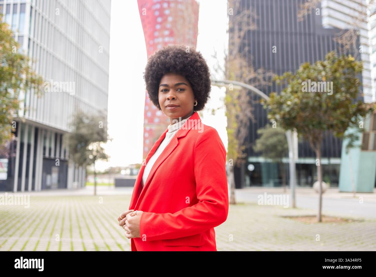Une femme confiante dans une veste rouge pose dans un cadre urbain moderne. Sa coiffure afro contraste magnifiquement avec l'arrière-plan architectural élégant. Banque D'Images