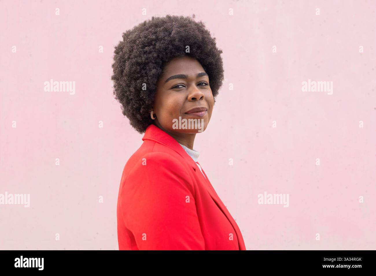 Une femme confiante dans un blazer rouge éclatant se dresse sur un fond rose doux, mettant en valeur l'élégance et la force. Sa coiffure afro ajoute une touche de pe Banque D'Images