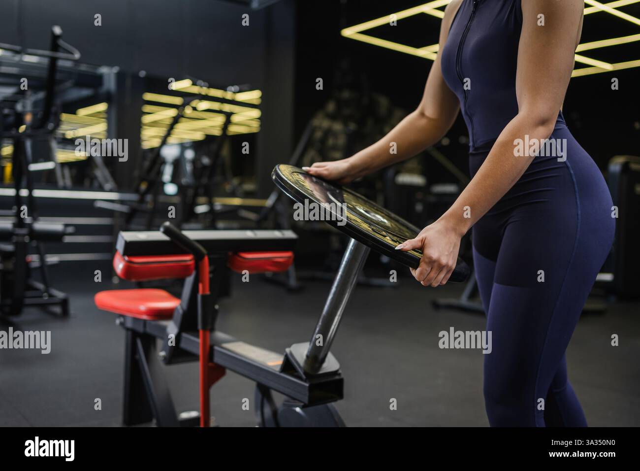 Une femme en sportswear sombre tient une plaque de poids dans un cadre de gymnastique moderne. Le centre de fitness est équipé d'appareils d'entraînement de pointe et d'un intérieur élégant Banque D'Images