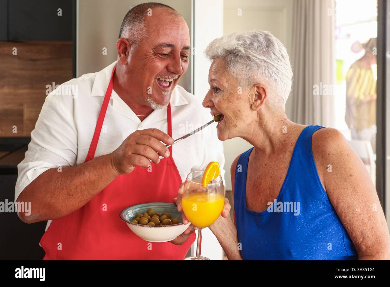 Un couple âgé joyeux s'engage dans une interaction ludique et affectueuse dans une cuisine lumineuse lors d'une fête, partageant des olives et du jus d'orange. Banque D'Images