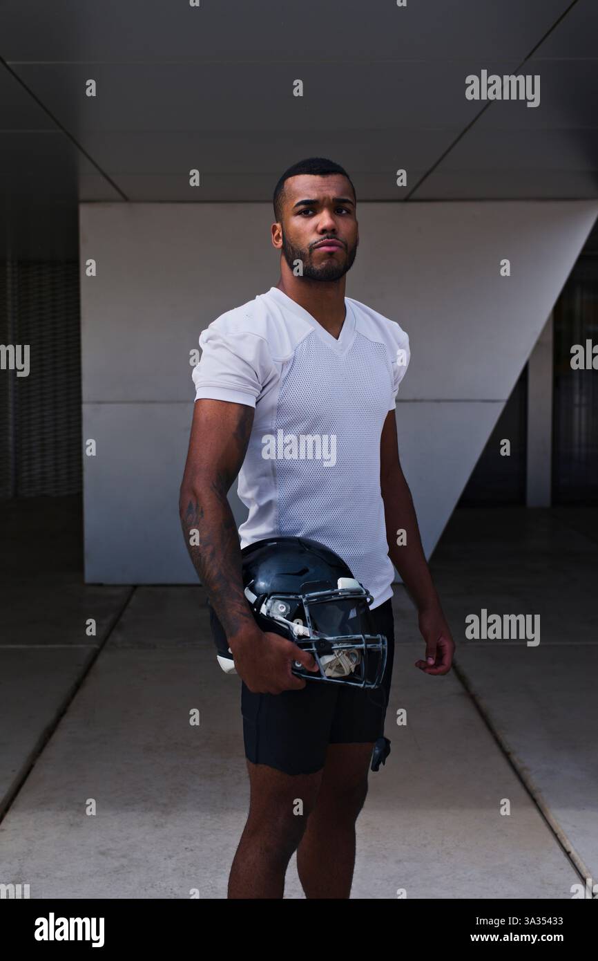 Un joueur de football américain confiant porte un maillot blanc, tenant un casque, prêt pour le match. L'athlète respire la détermination et la force. Banque D'Images