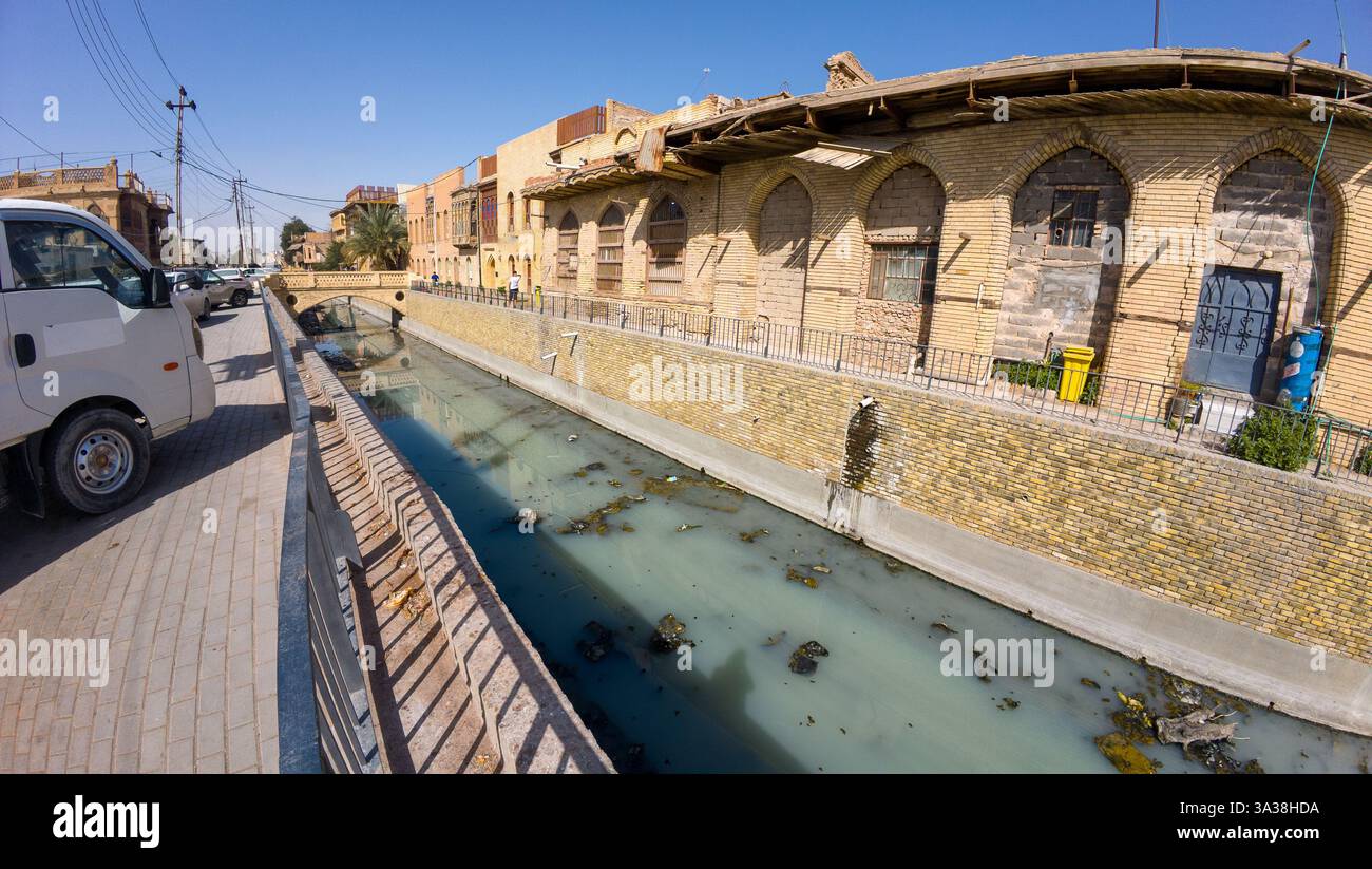 Un canal à l'eau trouble longe une rue avec des bâtiments en briques et un camion garé sous un ciel bleu clair. En Irak, Bassorah Banque D'Images