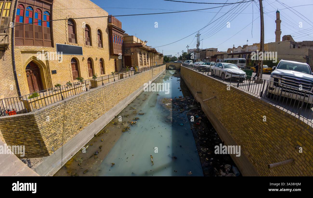 Canal urbain avec de l'eau trouble et des déchets, entouré de bâtiments en briques et de voitures garées. En Irak, Bassorah Banque D'Images