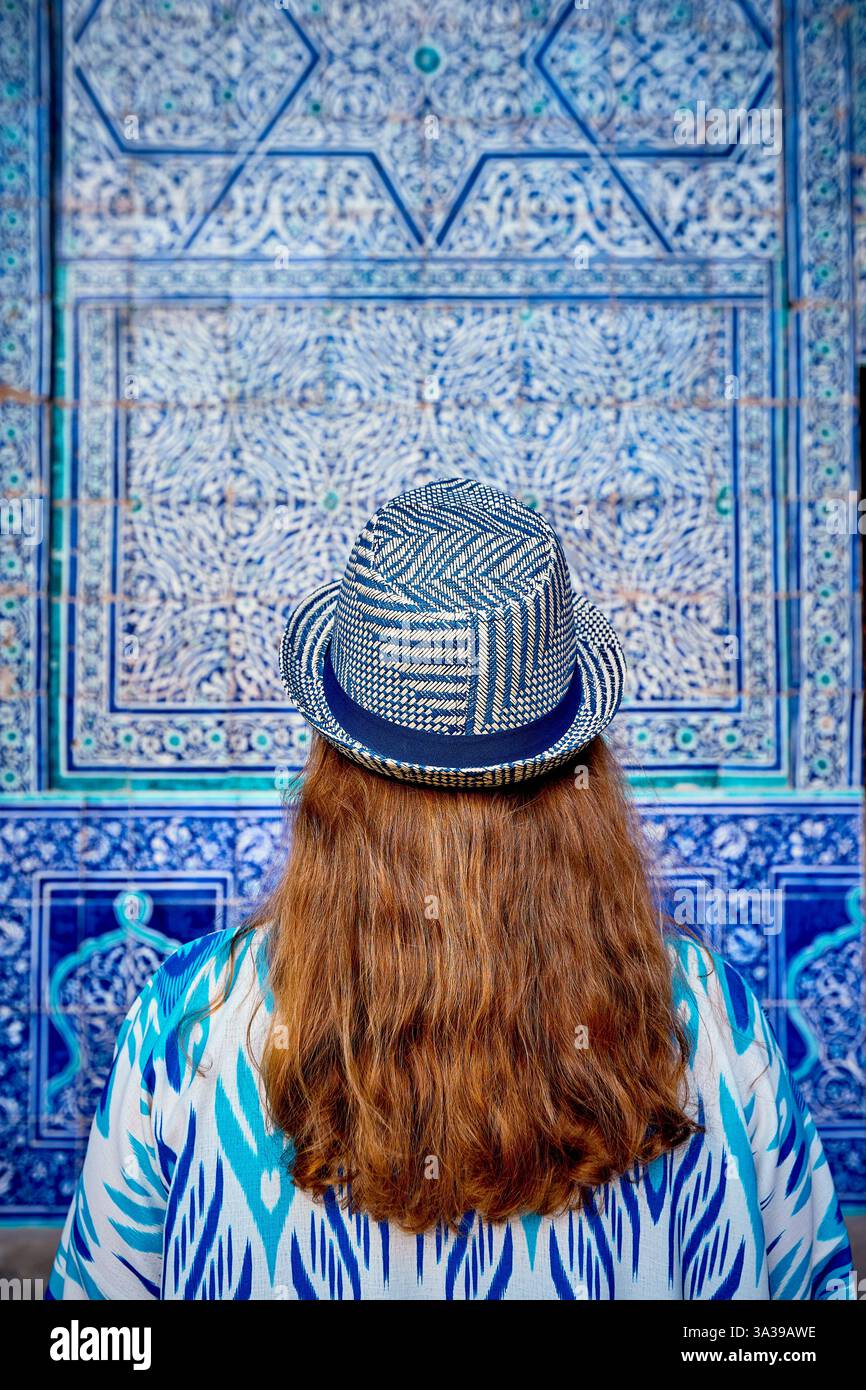 Femme touriste en robe ethnique et chapeau bleu devant le mur de mosaïque bleue dans le hall du palais à l'ancienne ville Khiva en Ouzbékistan Banque D'Images