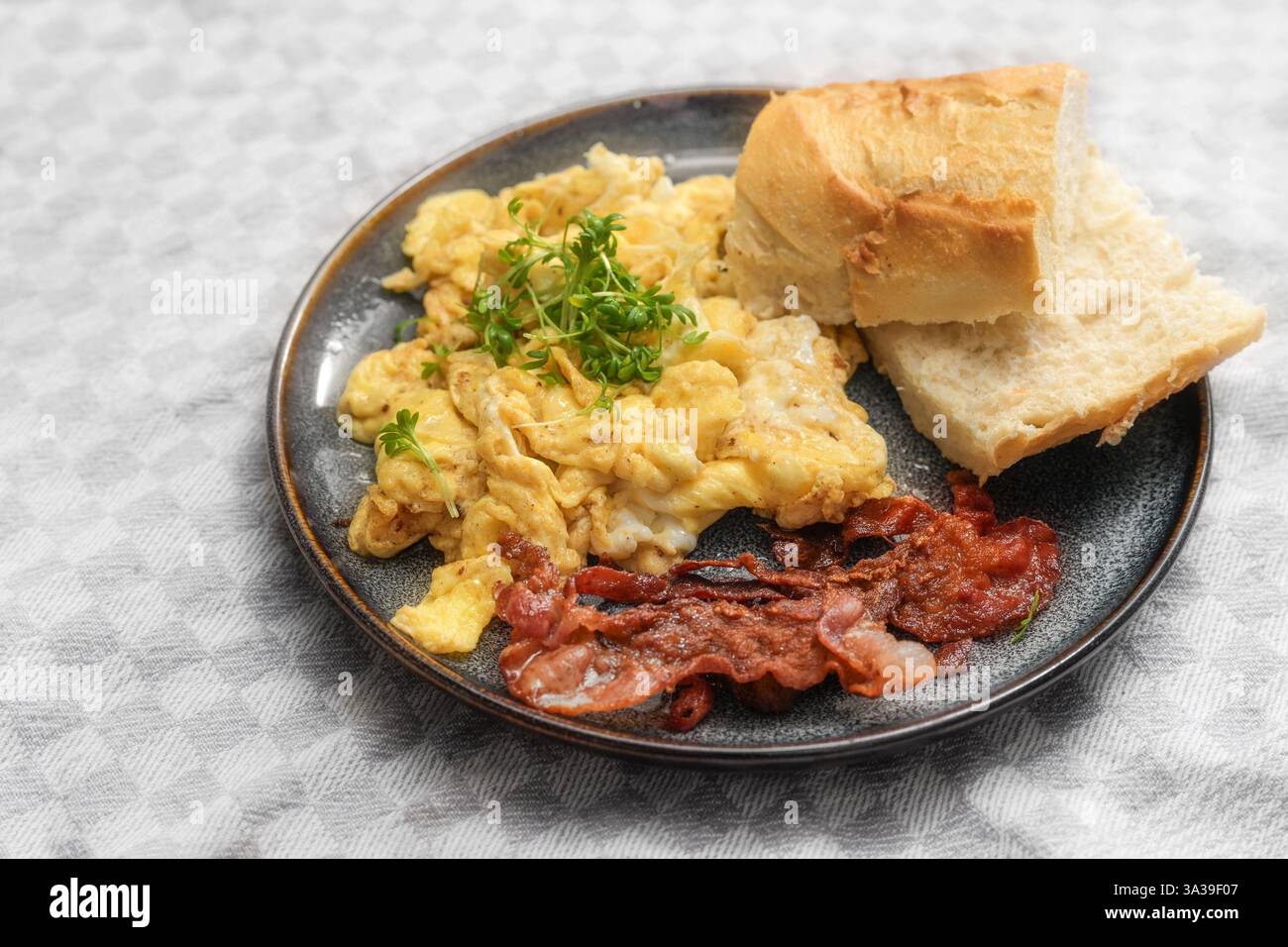 Œufs brouillés avec bacon croustillant, pain et garniture de cresson sur une assiette et une nappe gris clair, petit déjeuner copieux ou dîner rapide, focus sélectionné, na Banque D'Images