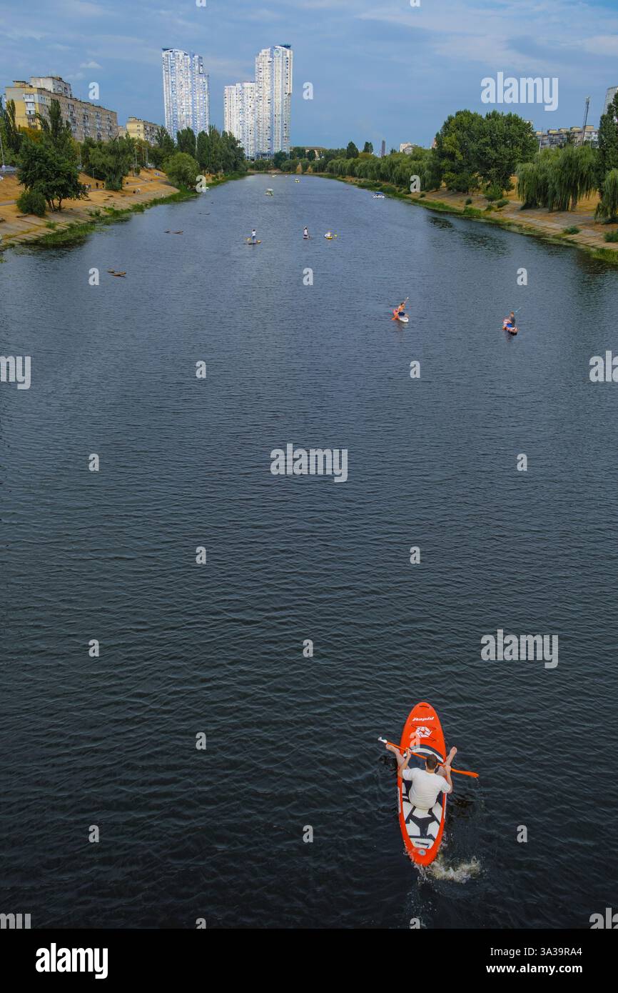Un groupe de kayakistes pagaie paisiblement le long d'une rivière tranquille, entourée d'une architecture urbaine et d'une végétation luxuriante par une journée lumineuse et ensoleillée. Banque D'Images