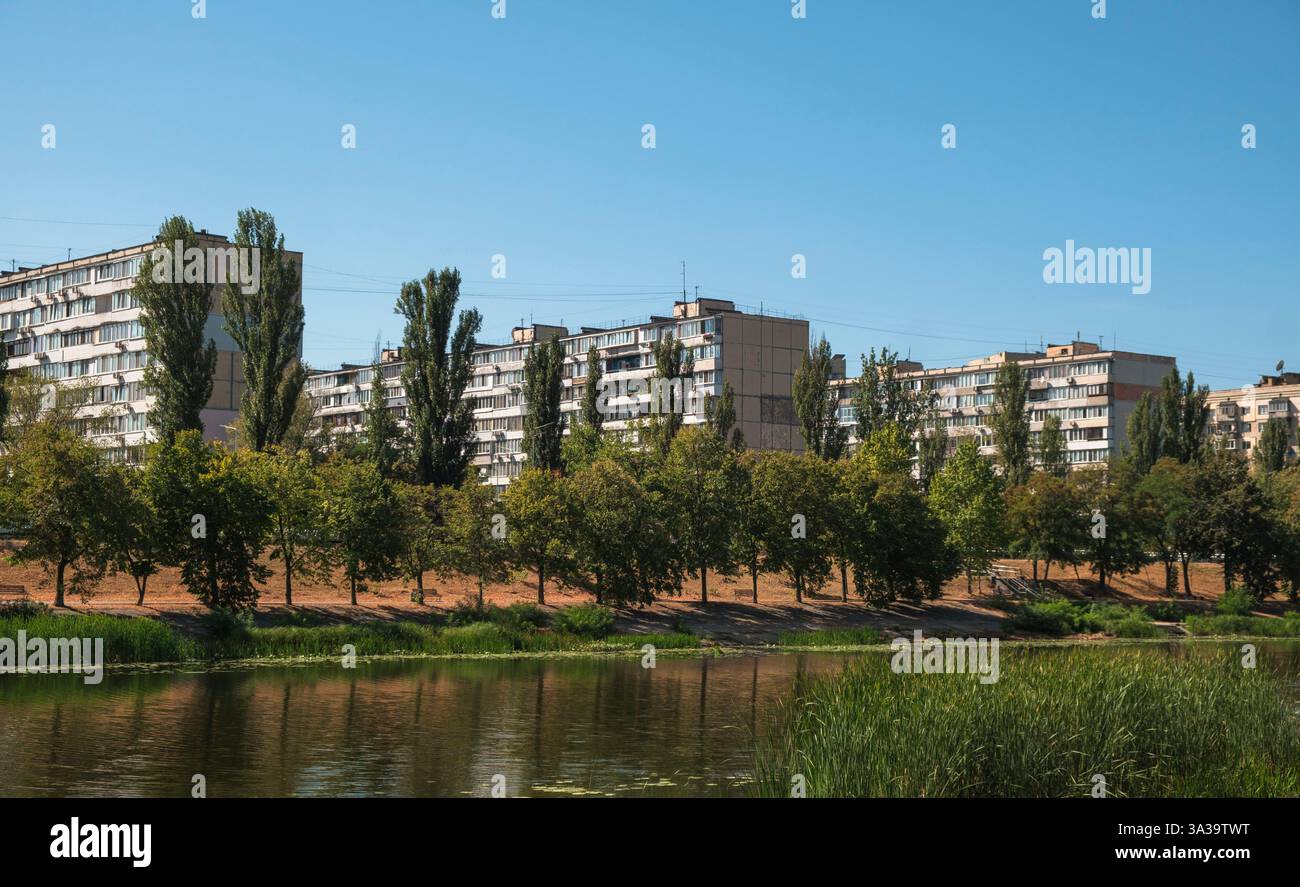 Les bâtiments modernes s'élèvent contre le ciel bleu clair tandis que les arbres luxuriants se reflètent dans les eaux calmes, créant une évasion sereine au bord de la rivière. Banque D'Images