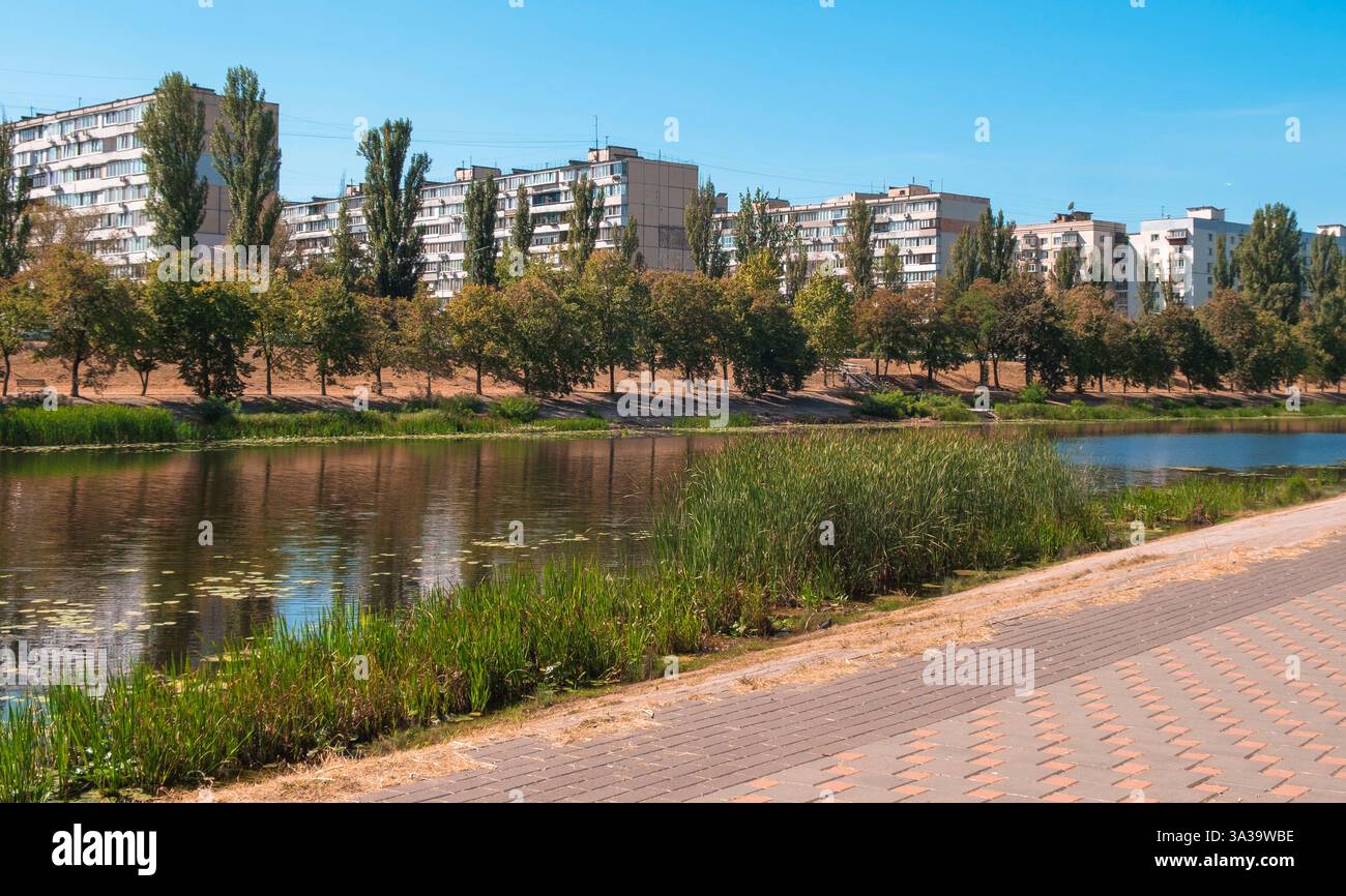 Un sentier paisible au bord de la rivière invite les visiteurs à profiter de la nature, avec des arbres et des bâtiments contemporains reflétant dans l'eau calme. Banque D'Images