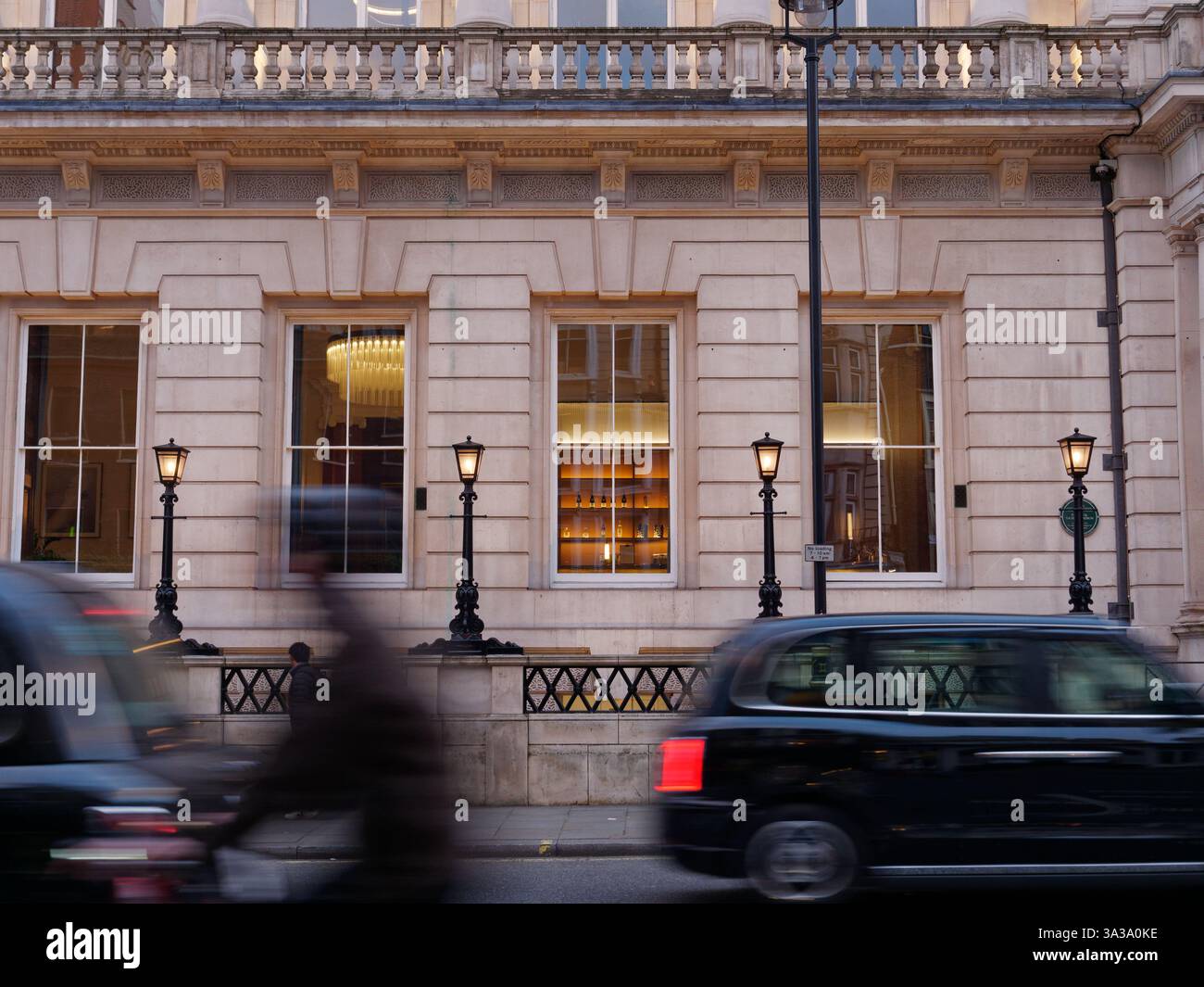 Taxi et cycliste en mouvement flou devant une façade de bâtiment ornée avec des lampadaires élégants allumés à Londres, mars 2025 Banque D'Images