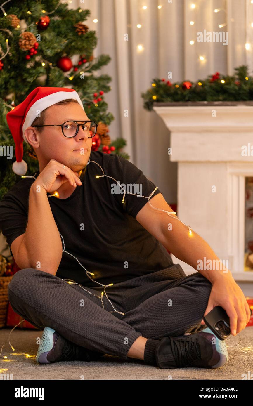 Un jeune gars est assis les jambes croisées dans des vêtements de maison et un chapeau de nouvel an sur fond d'un arbre de Noël et d'une cheminée Banque D'Images
