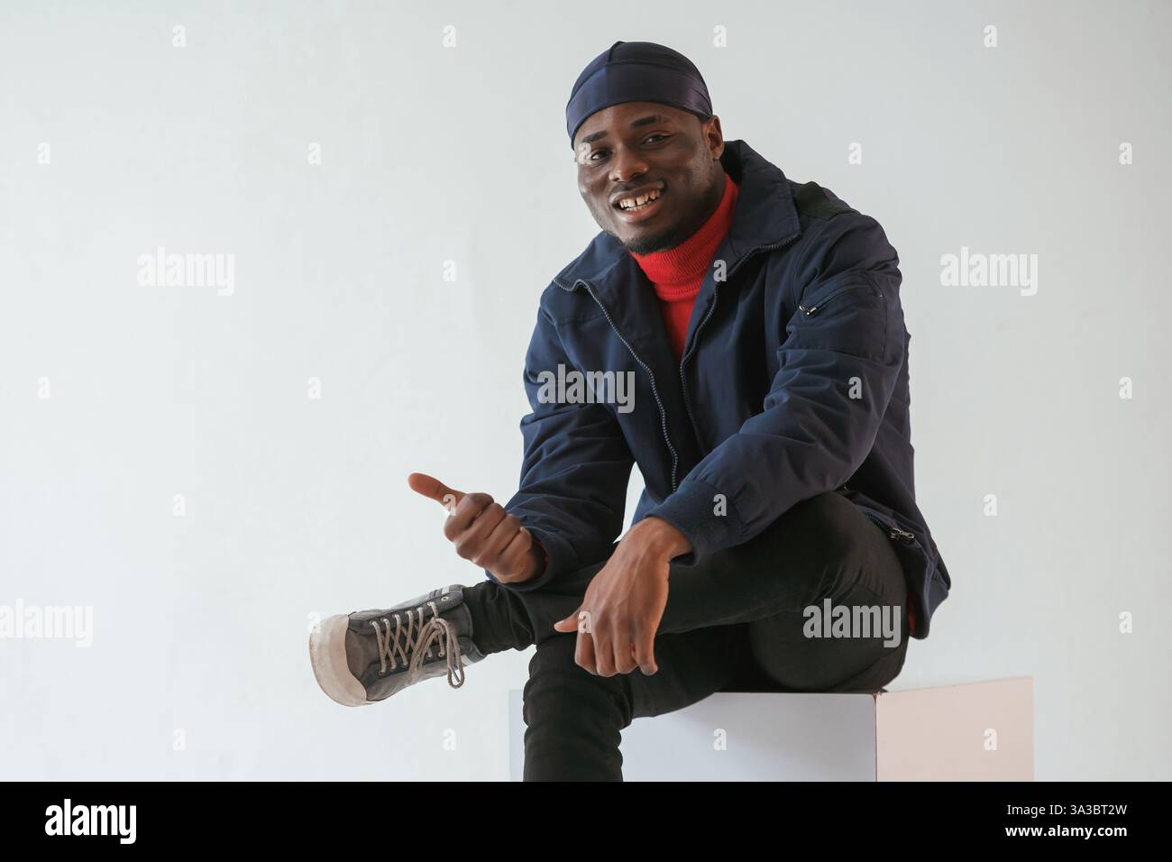 Calme, assis, souriant et montrant le pouce vers le haut. L'homme noir en casquette est dans le studio sur fond blanc. Banque D'Images