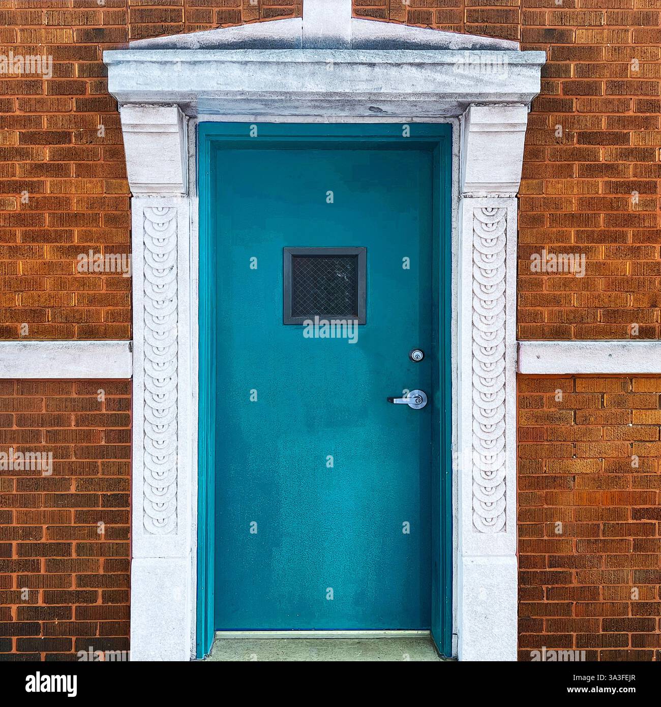Porte en métal sarcelle avec une petite fenêtre carrée, encadrée par une entrée en pierre blanche ornée, sertie contre une façade en briques brunes chaudes. Banque D'Images