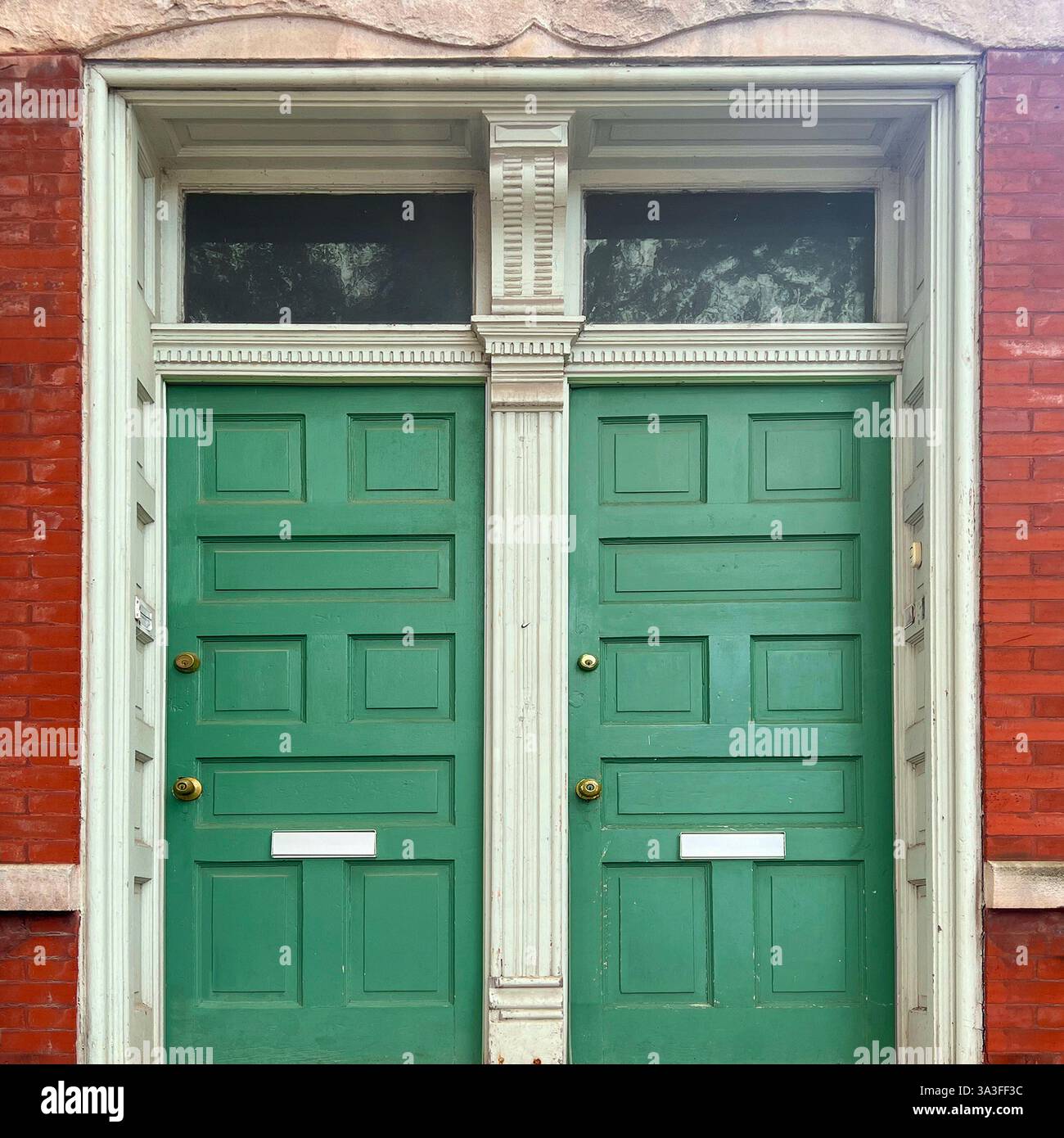 Portes doubles vertes vintage avec moulures décoratives blanches et fenêtres de tableau arrière, placées contre une façade historique en briques rouges. - Image de stock capturée avec un smartphone