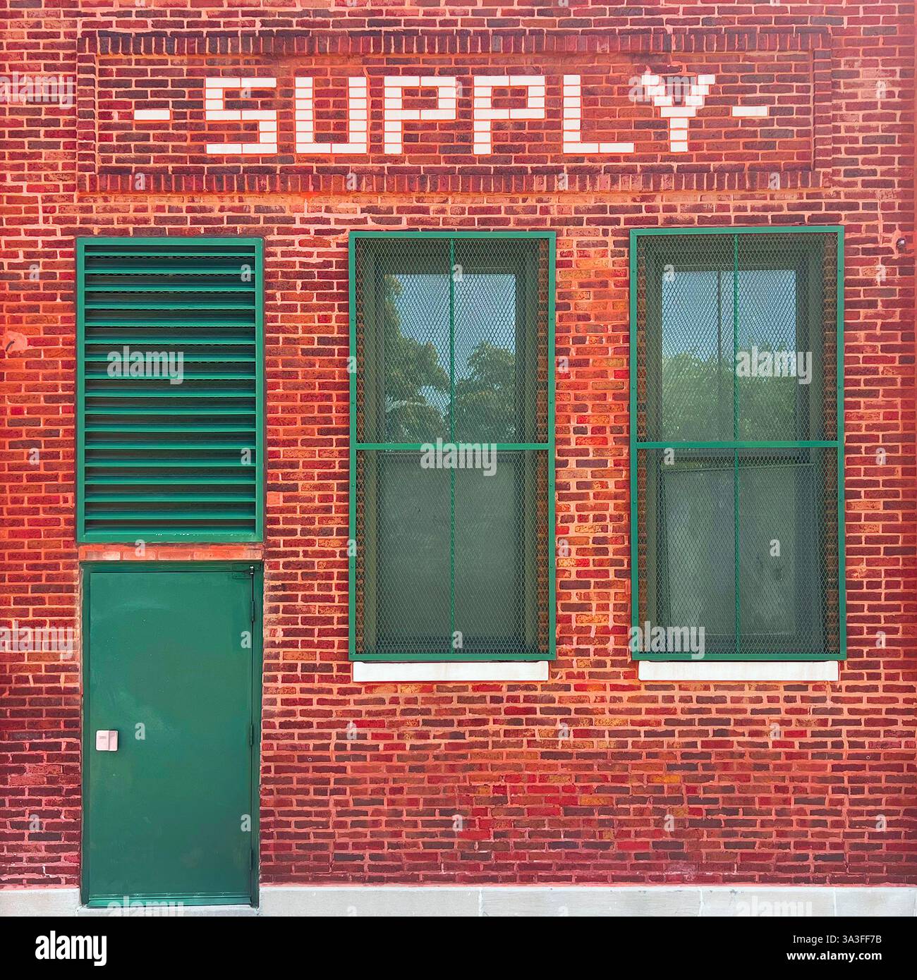 Un bâtiment historique d'approvisionnement en briques rouges avec une porte en métal vert, un panneau de ventilation et des fenêtres barrées de sécurité, avec une signalisation en lettres blanches audacieuses. Banque D'Images