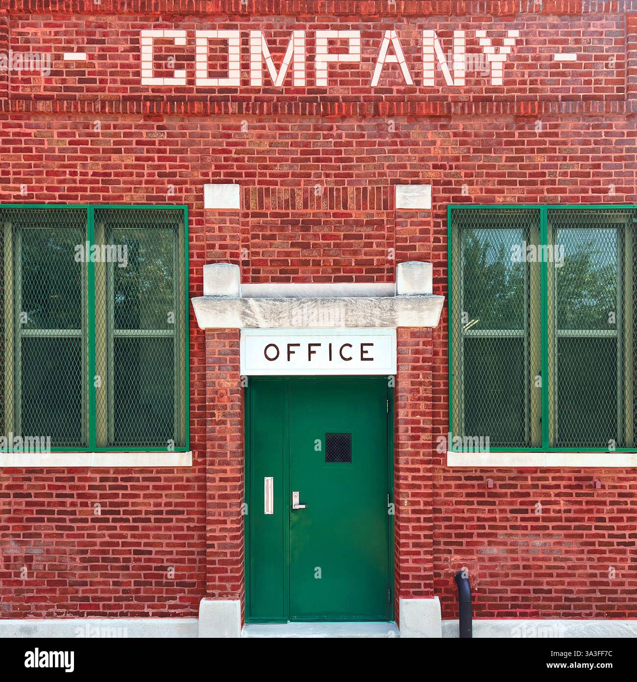 Un immeuble de bureaux en briques rouges vintage avec une porte en métal vert avec une petite fenêtre, encadrée par des fenêtres barrées de sécurité et une signalisation blanche audacieuse. Banque D'Images