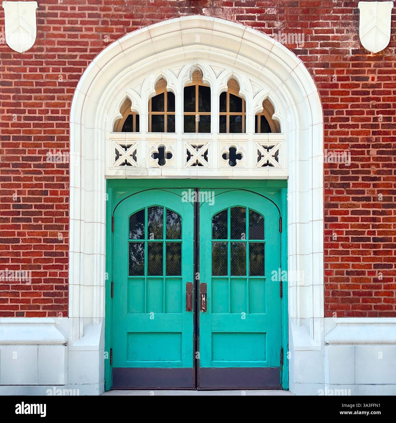 Double porte turquoise avec fenêtres cintrées, encadrée par des détails complexes en pierre de style gothique, située dans une façade en briques rouges. Banque D'Images