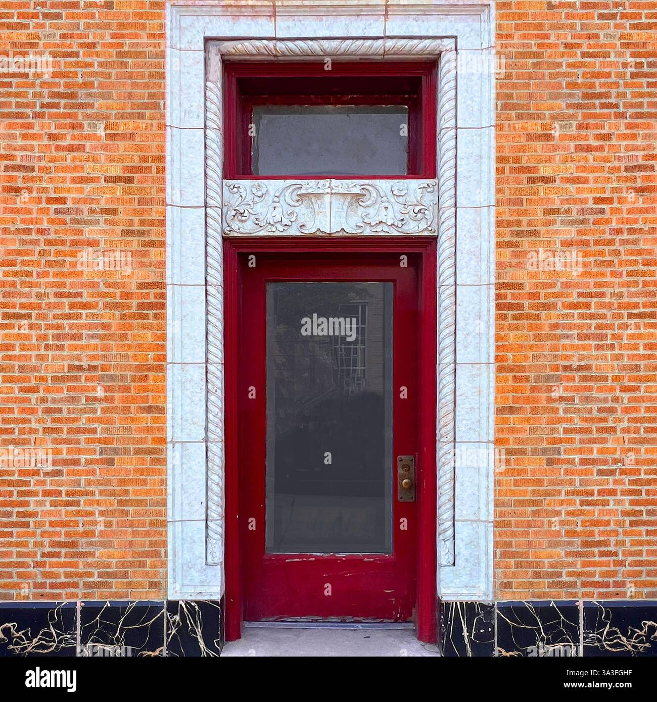 Porte rouge foncé avec panneau de verre et cadre en pierre blanche orné dans une façade en brique orange avec base décorative en marbre noir à Chicago. - Image de stock capturée avec un smartphone