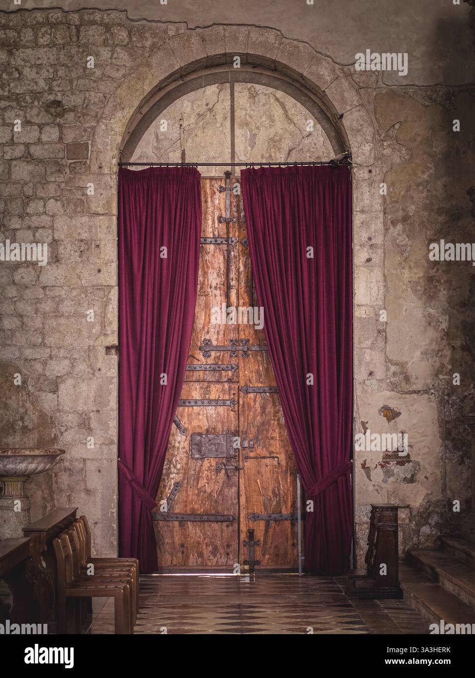 Une porte en bois vieilli avec des détails en fer, encadrée par des rideaux de velours rouge profond dans un vieux bâtiment en pierre. Les murs vieillis et l'éclairage chaleureux évoquent un sens Banque D'Images